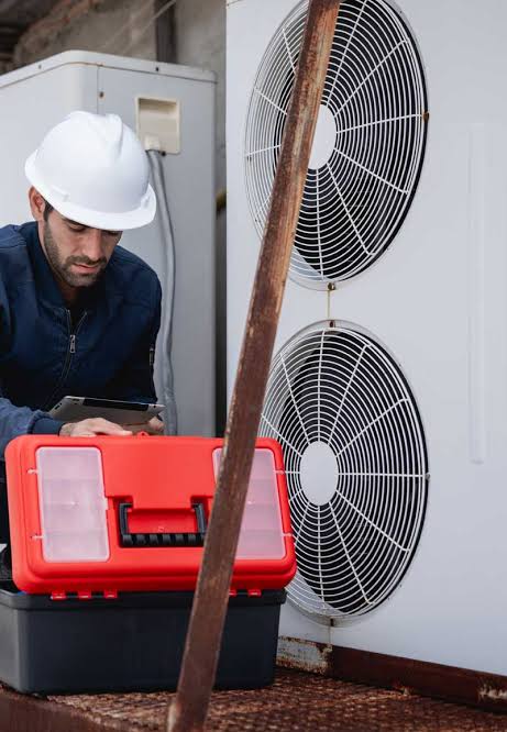 Technician in a white hard hat inspecting an outdoor HVAC unit beside a red toolbox